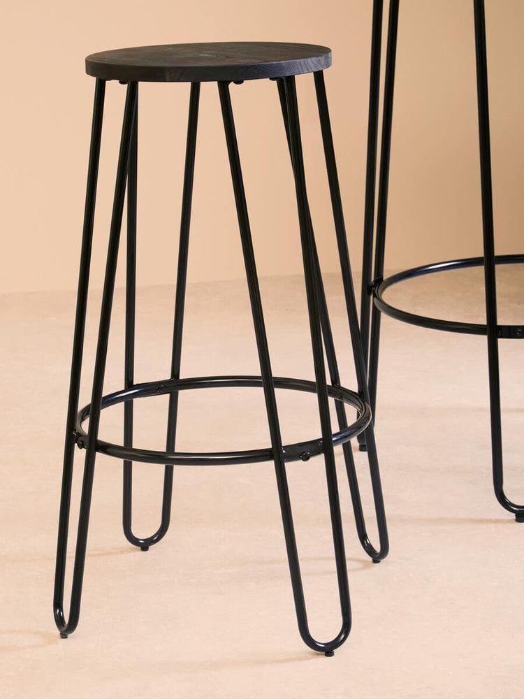 A black metal bar stool with a round seat and slender legs, featuring a minimalist design. The stool is positioned next to a tall bar table, showcasing the set's modern aesthetic against a neutral background