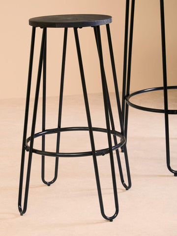 A black metal bar stool with a round seat and slender legs, featuring a minimalist design. The stool is positioned next to a tall bar table, showcasing the set's modern aesthetic against a neutral background