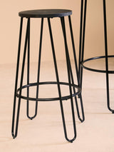 A black metal bar stool with a round seat and slender legs, featuring a minimalist design. The stool is positioned next to a tall bar table, showcasing the set's modern aesthetic against a neutral background