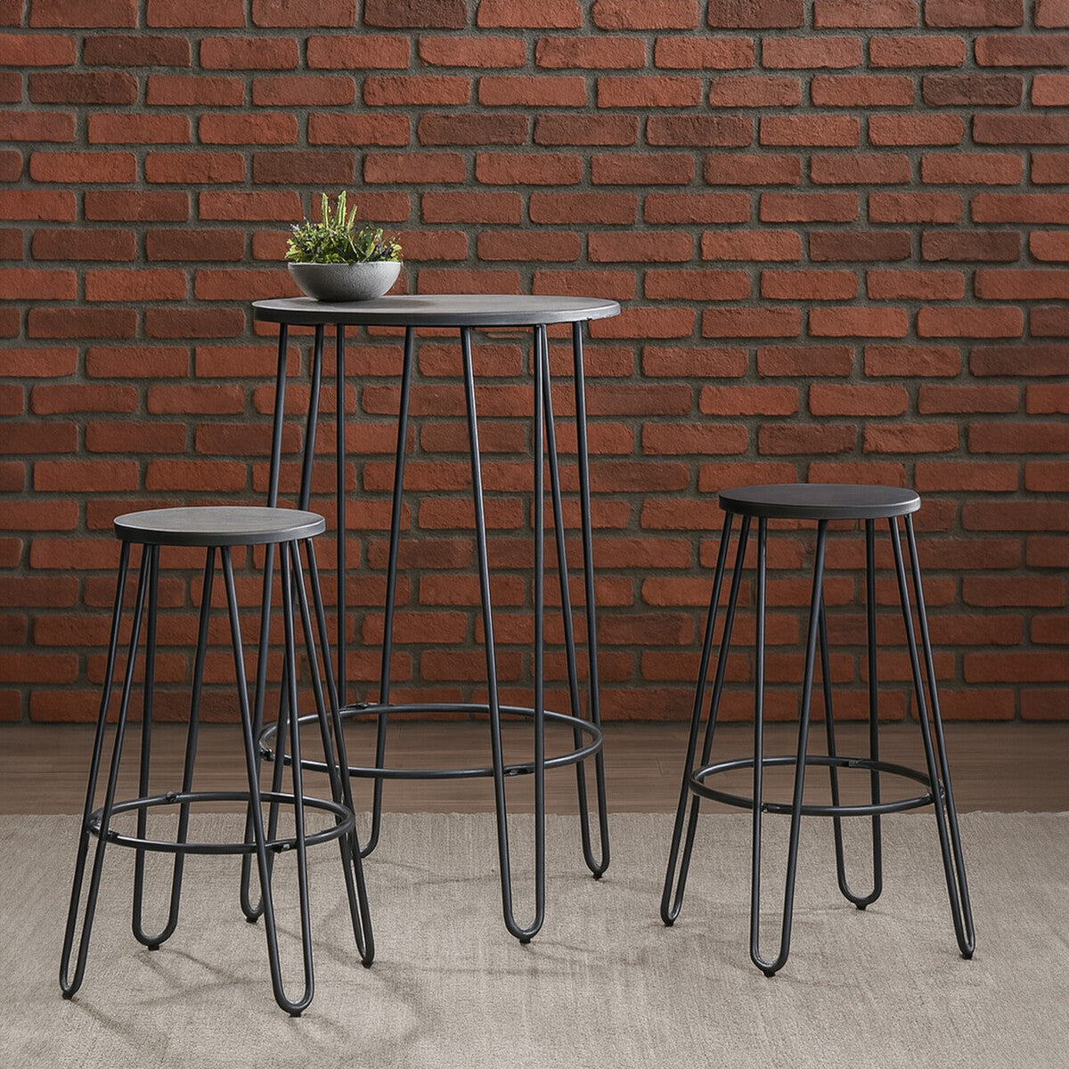 A three-piece bar table and stool set featuring a tall, rectangular table with a smooth surface and sleek black metal legs, accompanied by two matching stools with round seats and sturdy bases. The background consists of a textured brick wall, and a small potted plant sits on the table