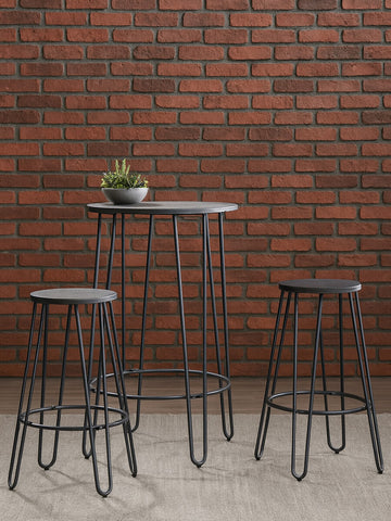 A three-piece bar table and stool set featuring a tall, rectangular table with a sleek black surface and slender metal legs, accompanied by two matching bar stools with round seats and similar metal legs. The set is positioned against a textured brick wall, creating a modern and stylish look. A small potted plant is placed on the table for added decor