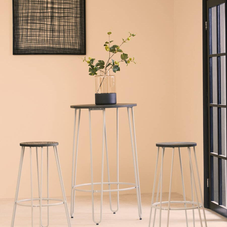 Three-piece bar table and stool set featuring a sleek elm wood tabletop and two matching stools with wooden seats. The table stands on slender metal legs, and a small potted plant is placed on the table. The background includes a light-colored wall and a window with black frames, creating a modern and inviting atmosphere