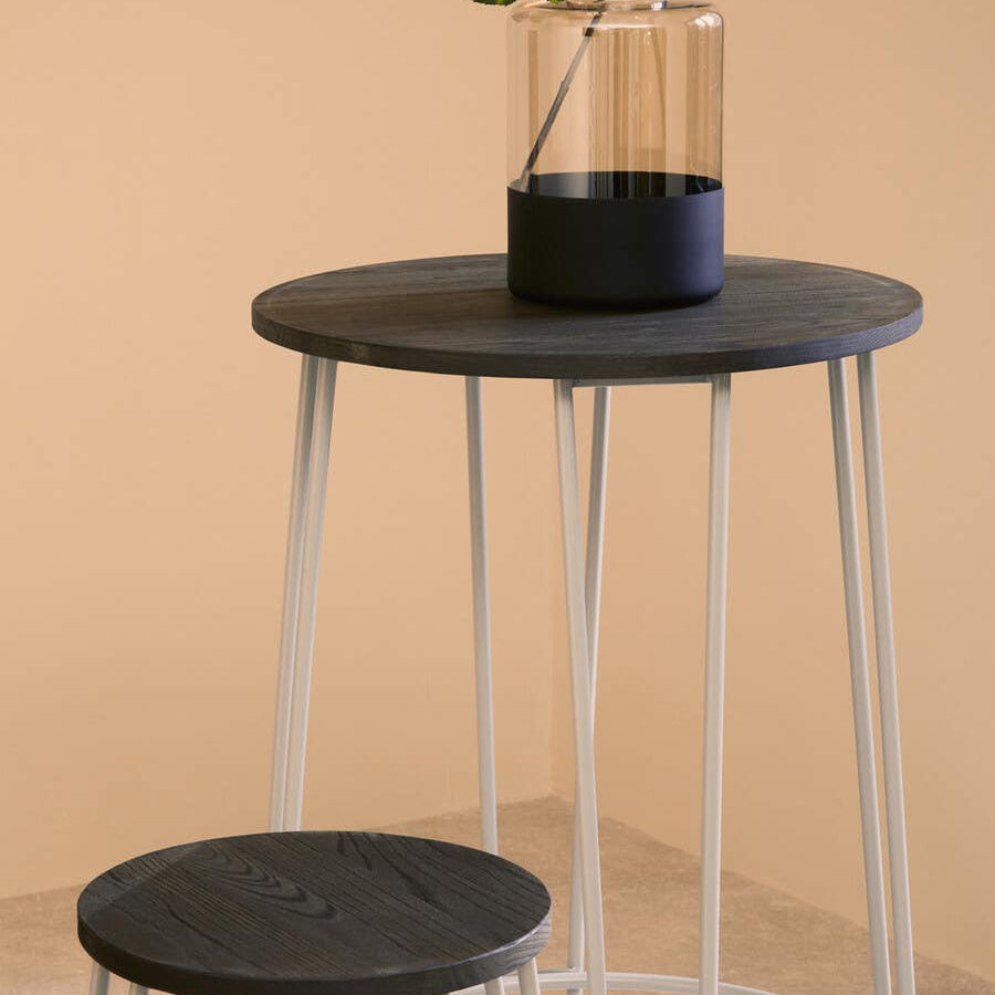 A round black elm wood bar table with slender metal legs, accompanied by a matching stool. The table features a glass vase with greenery on top, set against a soft beige background