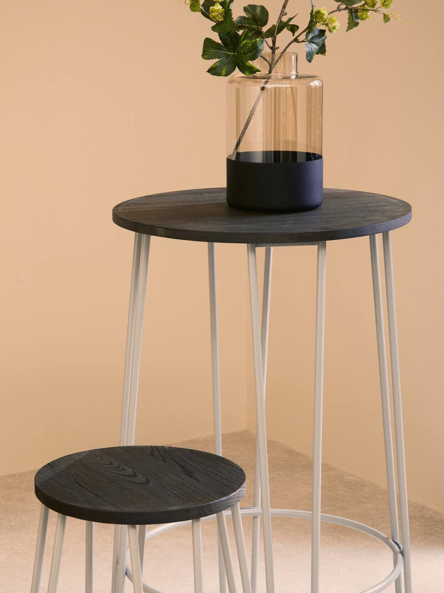 A round black elm wood bar table with slender metal legs, accompanied by a matching stool. The table features a glass vase with greenery on top, set against a soft beige background
