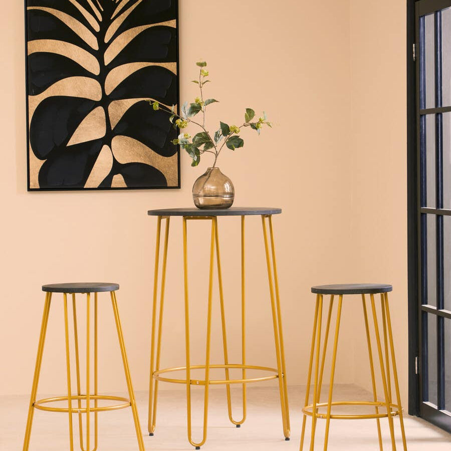A three-piece bar table and stool set featuring a gold finish with sleek, modern design. The set includes one tall bar table and two matching stools, all with black tops. The background showcases a minimalist wall art piece with a plant design and a light-colored wall, creating a stylish and contemporary atmosphere