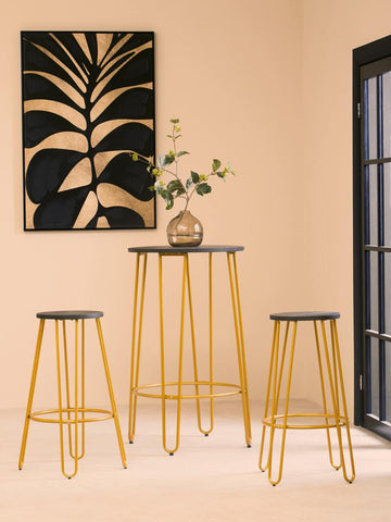 A three-piece bar table and stool set featuring a gold finish with sleek, modern design. The set includes one tall bar table and two matching stools, all with black tops. The background showcases a minimalist wall art piece with a plant design and a light-colored wall, creating a stylish and contemporary atmosphere