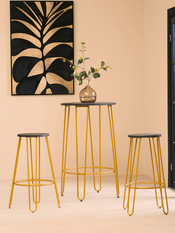 Three-piece bar table and stool set featuring a sleek gold finish. The set includes a tall bar table with a round top and two matching stools with round seats. The background showcases a light-colored wall and a decorative wall art piece with a plant motif. A small vase with greenery is placed on the table