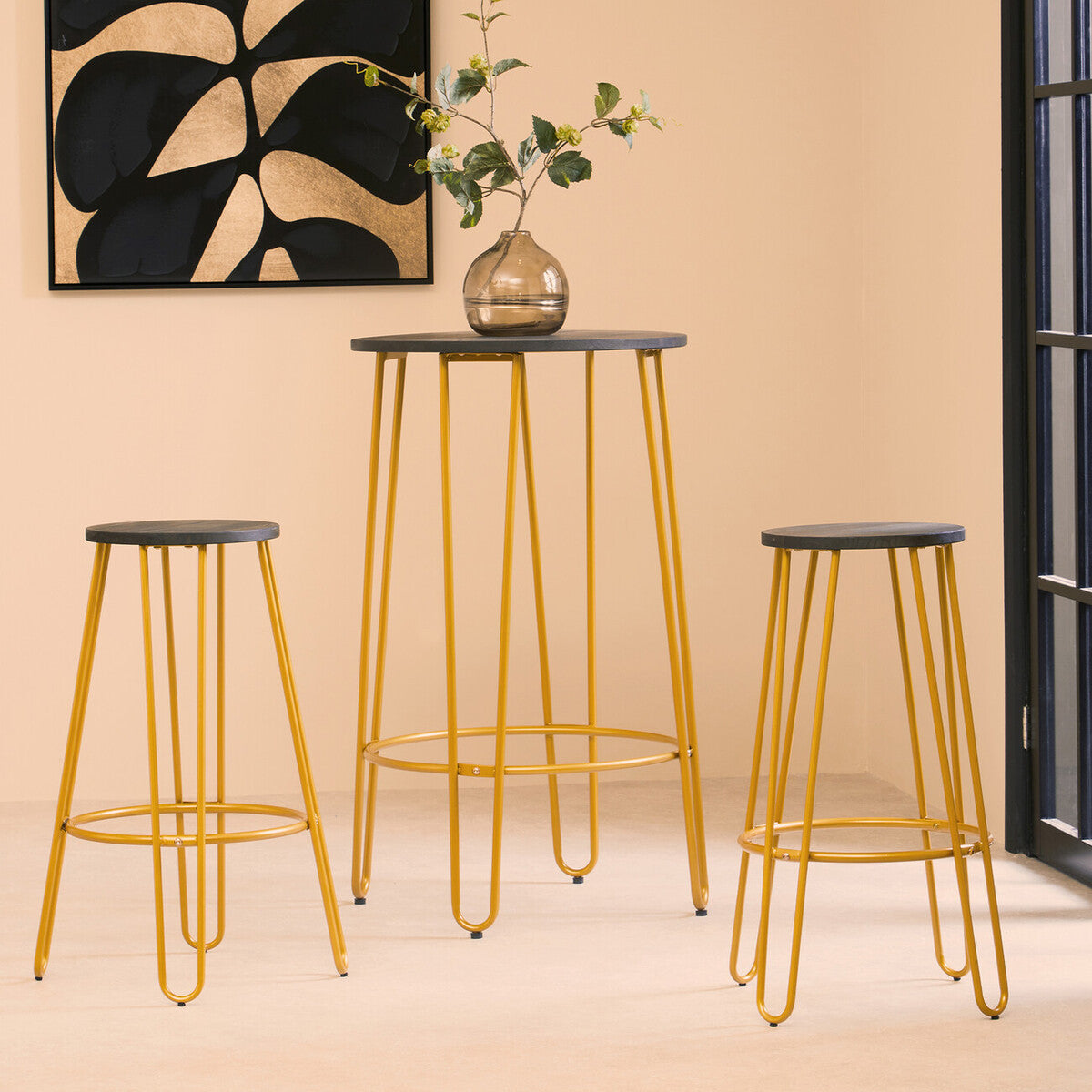 A three-piece bar table and stool set featuring a sleek gold finish. The set includes a tall bar table with a round top and three matching stools with a minimalist design. The table and stools have slender, elegant legs, and the background showcases a neutral-colored wall with a decorative plant and a window
