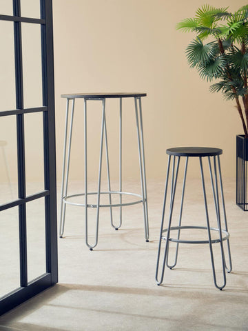 Two bar stools with a grey finish metal frame are positioned in a bright room. One stool is taller, while the other is shorter, both featuring a simple, modern design. A potted plant is visible in the background, and a glass door frame is partially shown on the left side. The floor is light-colored, enhancing the airy feel of the space