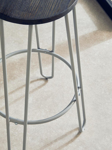 A grey metal frame bar stool with a round wooden seat, featuring four slender legs and a circular footrest, set against a light-colored floor