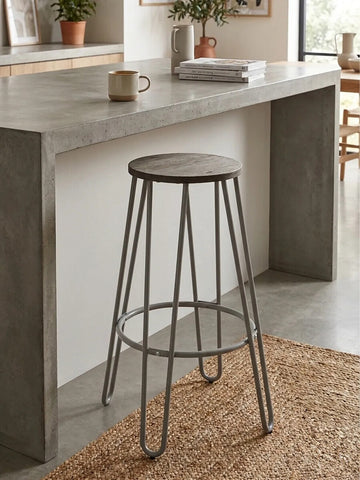 A grey finish metal frame bar stool with a simple, modern design, positioned next to a concrete countertop. The stool features a round seat and slender legs, with a circular footrest. A woven rug is visible on the floor beneath the stool, adding texture to the scene