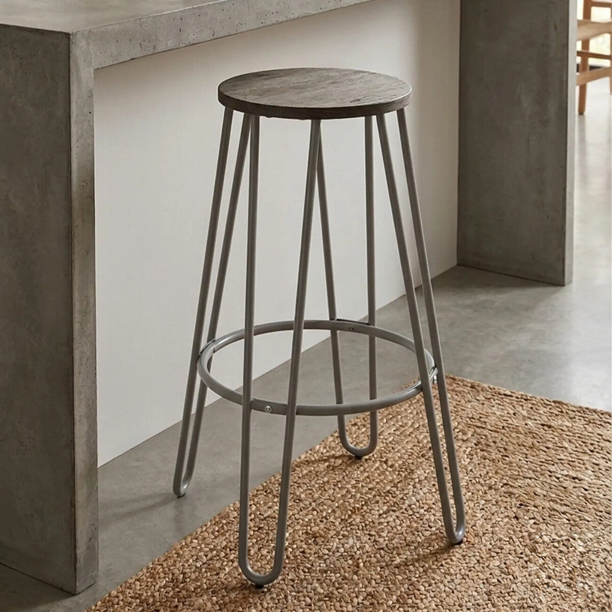 A grey finish metal frame bar stool with a round wooden seat, featuring slender legs and a footrest, positioned next to a concrete bar counter on a textured rug