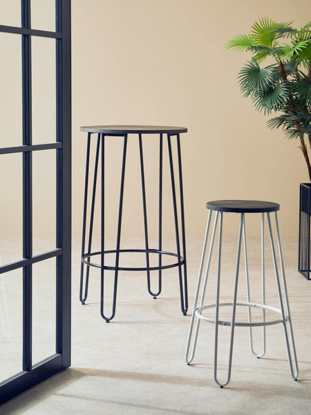Two bar stools with a white finish metal frame are positioned in a bright room. One stool is taller, while the other is shorter, both featuring a simple, modern design. A green plant is visible in the background, and the floor has a light-colored finish. A black-framed window is partially visible on the left side of the image