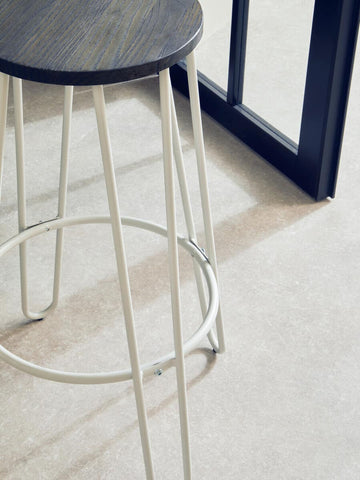 A white finish metal frame bar stool with a round seat and slender legs, positioned on a light-colored floor next to a black-framed structure. The stool features a circular footrest and a minimalist design, emphasizing modern aesthetics