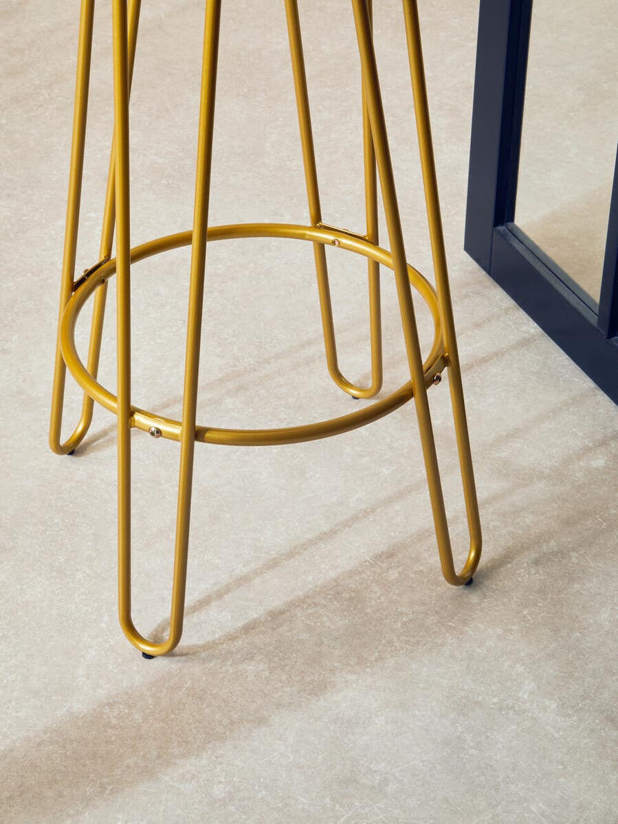 Gold finish metal frame bar stool with slender legs and a circular footrest, set against a light-colored floor and a dark blue structure in the background