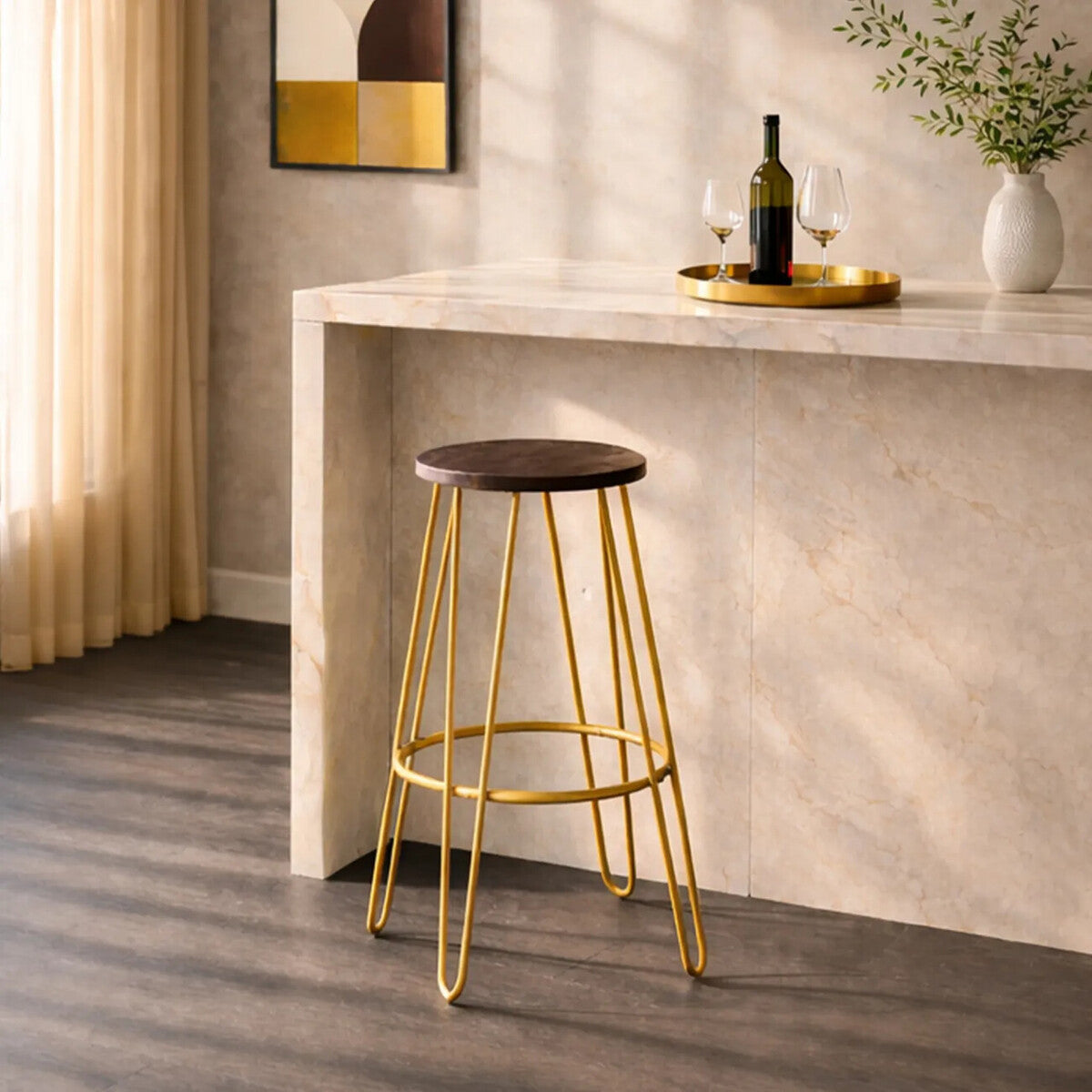 Gold finish metal frame bar stool with a round wooden seat, positioned next to a light-colored countertop. Soft natural light filters through sheer curtains, highlighting the stool's sleek design and the warm tones of the wood and metal. A bottle of wine and a glass are visible on the countertop, adding to the inviting atmosphere