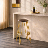 Gold finish metal frame bar stool with a round wooden seat, positioned next to a light-colored countertop. Soft natural light filters through sheer curtains, highlighting the stool's sleek design and the warm tones of the wood and metal. A bottle of wine and a glass are visible on the countertop, adding to the inviting atmosphere
