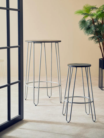 Two District Barstools with silver metal legs are positioned in a bright space, one taller and one shorter, against a light-colored wall. A potted plant is visible in the background, adding a touch of greenery to the scene. The floor appears to be light-colored wood or laminate