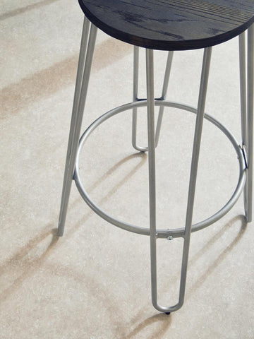 A barstool featuring a round wooden seat with a dark finish and sleek silver metal legs, showcasing a minimalist design. The legs are thin and tapered, with a circular footrest connecting them, set against a light-colored floor