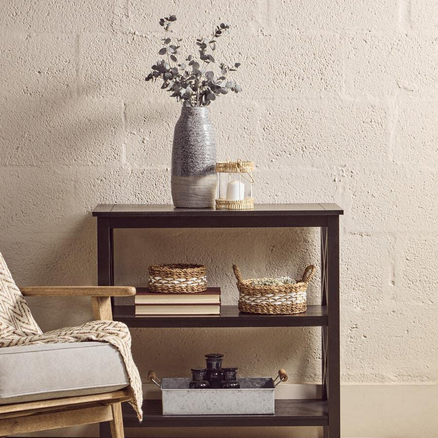 Dark wood console table with shelves, baskets, books, ceramic vase and candle holder beside chair