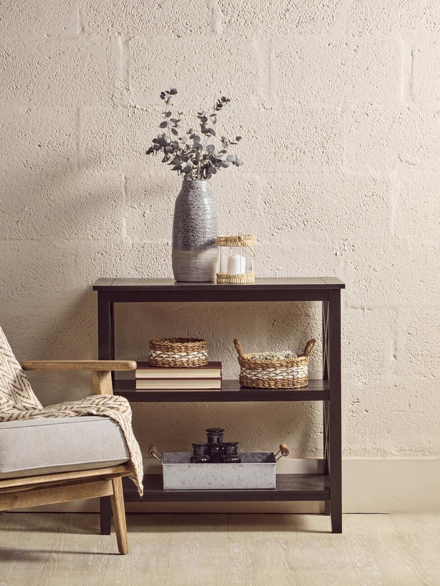 Dark wood console table with shelves, baskets, books, ceramic vase and candle holder beside chair