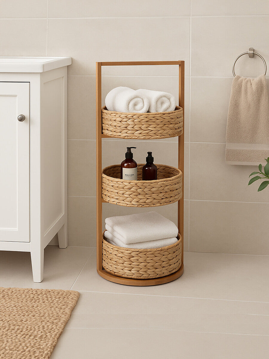 Three-tier storage caddy made from water hyacinth with a natural wood frame, featuring two open shelves and a top handle. The caddy holds neatly stacked white towels and two dark bottles, positioned next to a white cabinet in a softly lit room with neutral-colored walls and a light-colored floor