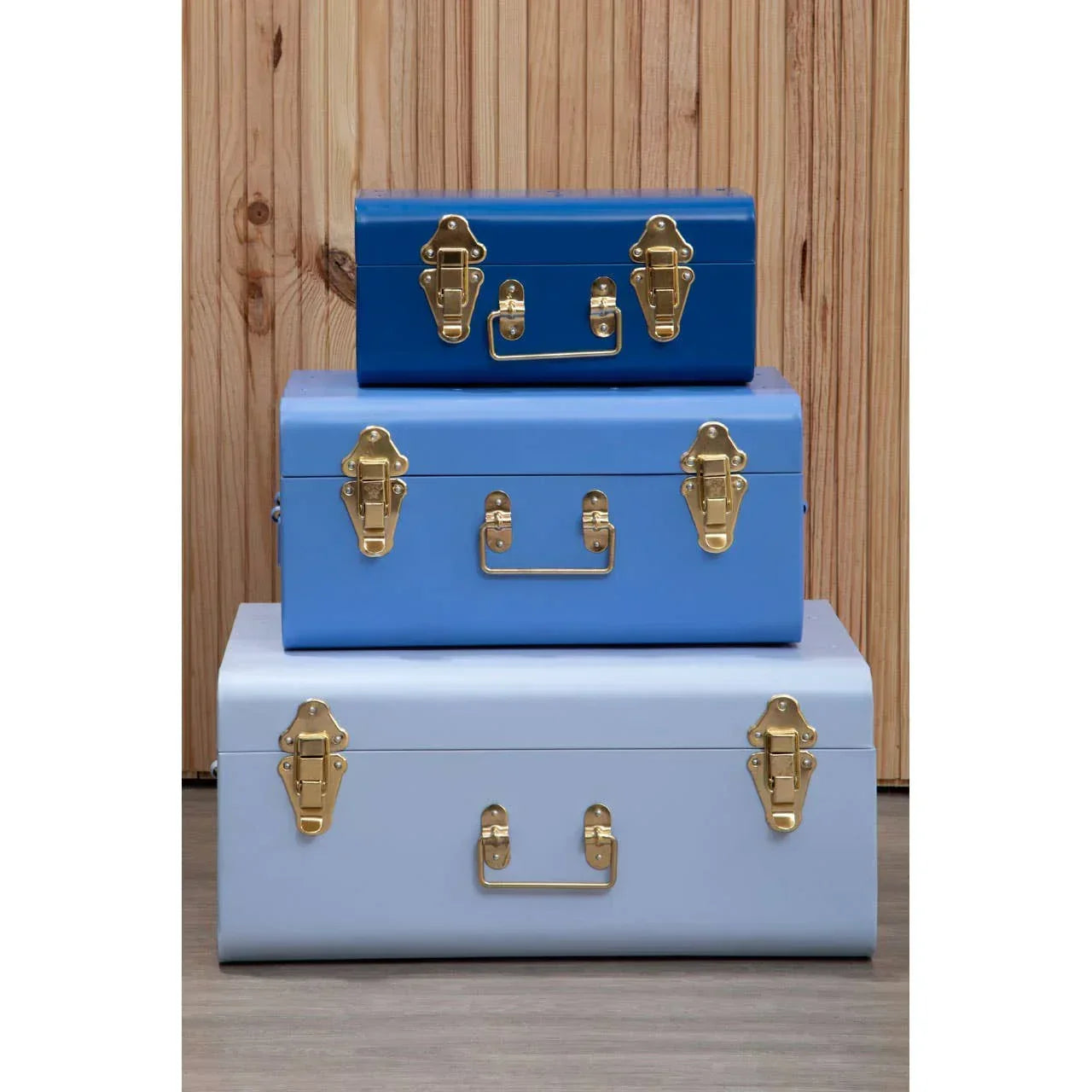 Set of three blue metal storage trunks with gold latches stacked against wooden panel wall