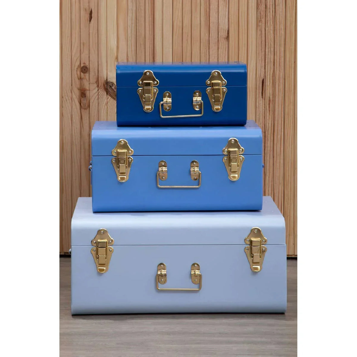 Set of three blue metal storage trunks with gold latches stacked against wooden panel wall