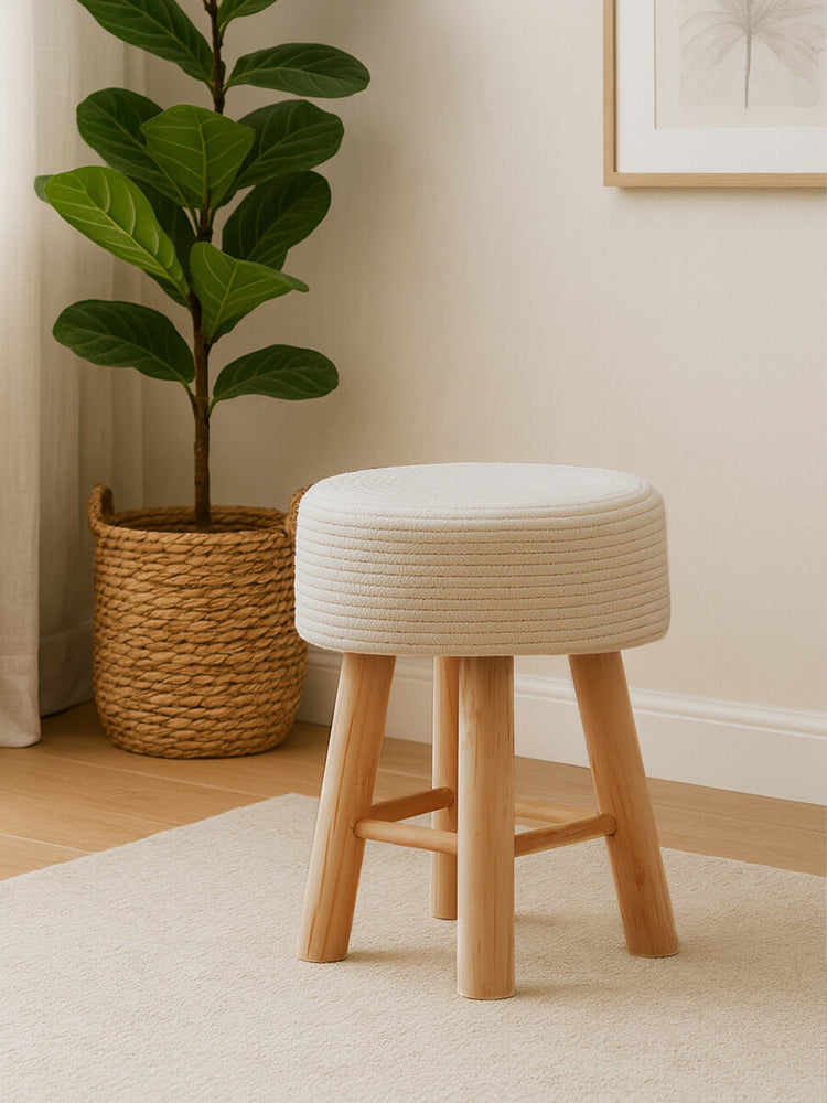 A white cotton rope stool with a round top and natural wood legs, placed on a light-colored floor next to a potted plant in a woven basket. The background features a neutral wall and a hint of artwork