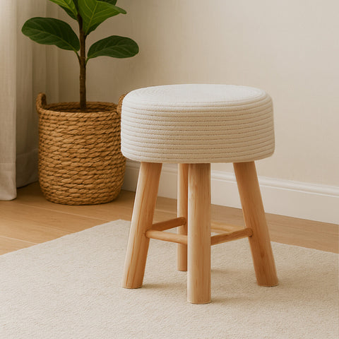 A white cotton rope stool with a round top and natural wooden legs, placed on a light-colored rug in a cozy room setting, with a woven basket and a green plant in the background