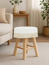 A natural wood stool with a white cushioned top, featuring four wooden legs. The stool is positioned next to a light-colored sofa in a cozy living room setting, with a potted plant and decorative items visible in the background