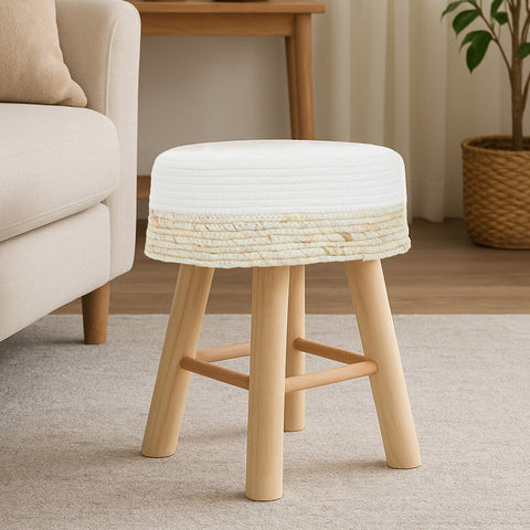 A natural wood stool with a white cushioned top and four light wooden legs, placed on a beige carpet in a cozy living room setting. A sofa and a plant are visible in the background
