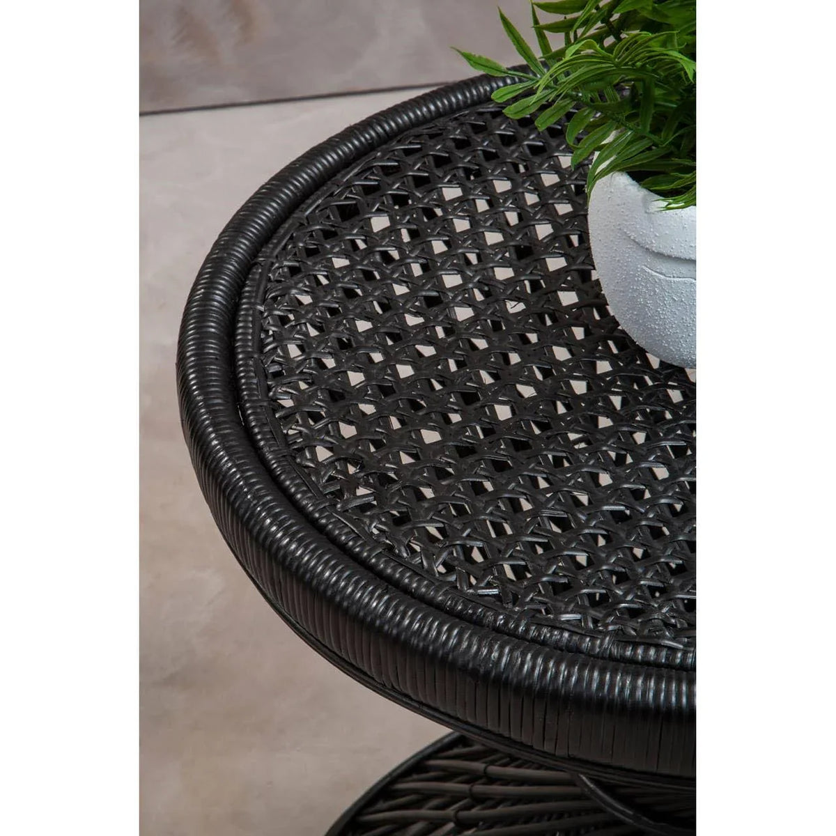 Black rattan side table with woven top and potted plant on light stone floor
