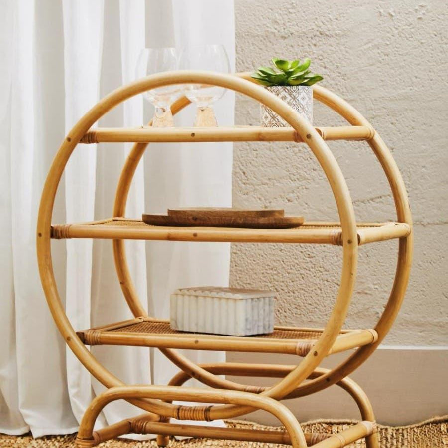 Round rattan side table with three shelves holding glassware, plant, and decor accents