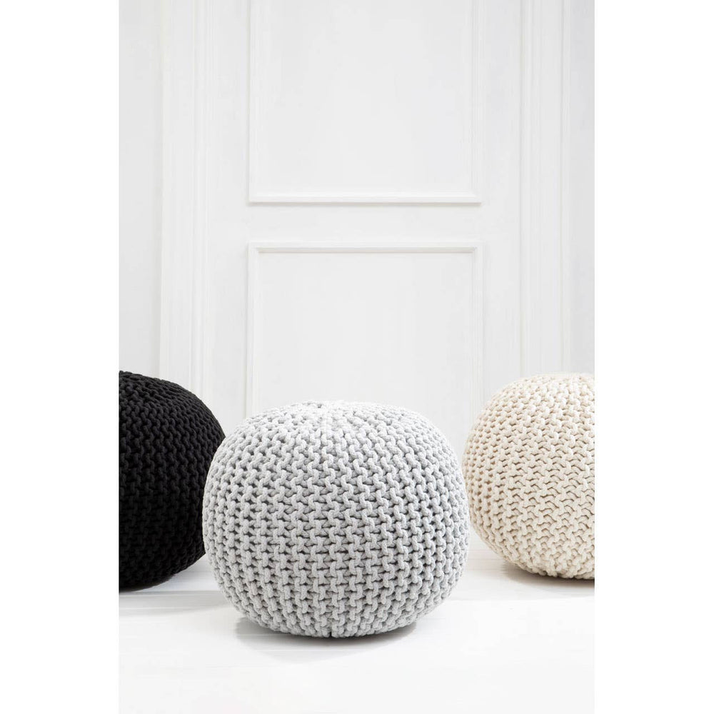 A grey knit pouffe with a textured surface, positioned among a black pouffe and a beige pouffe, set against a white wall with a door in the background