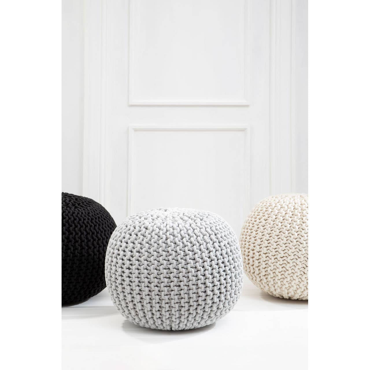 A grey knit pouffe with a textured surface, positioned among a black pouffe and a beige pouffe, set against a white wall with a door in the background