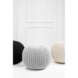 A grey knit pouffe with a textured surface, positioned among a black pouffe and a beige pouffe, set against a white wall with a door in the background