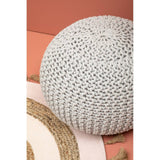 A round grey knit pouffe resting on a textured pink and beige surface, showcasing its intricate knitted pattern and soft, cozy appearance