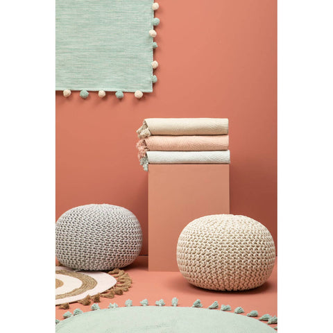 A grey knit pouffe sits on a textured rug alongside another pouffe, with a stack of folded blankets in soft pastel colors on a pedestal. The background features a coral wall and a light green rug with a scalloped edge, creating a cozy and inviting atmosphere