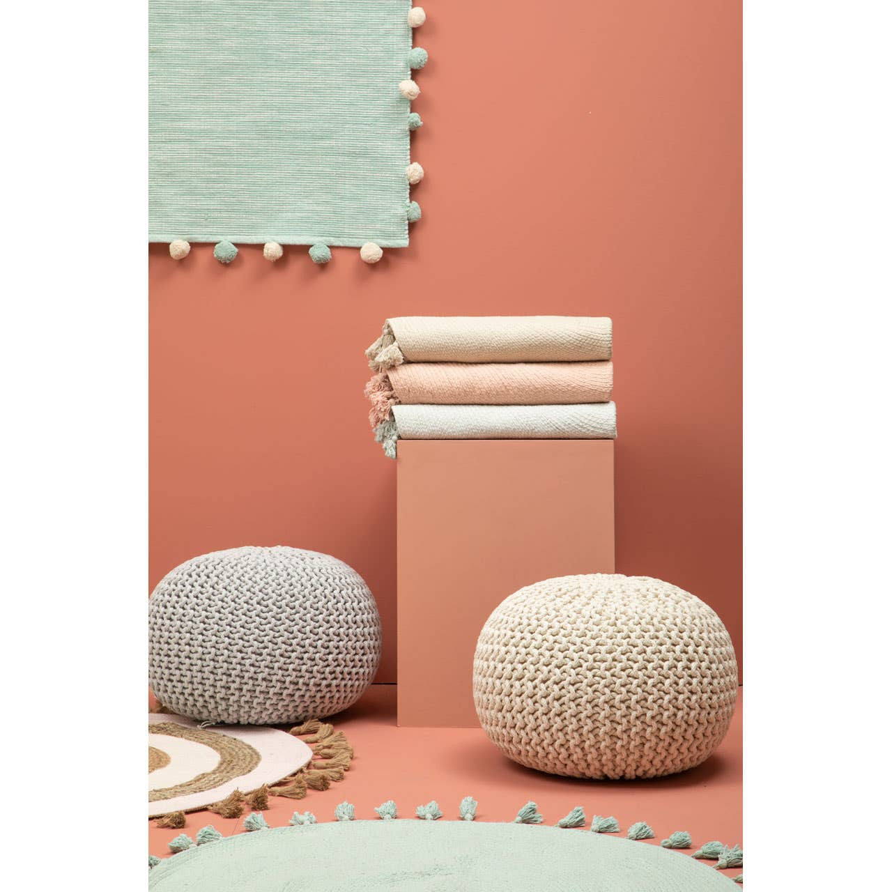 A grey knit pouffe sits on a textured rug alongside another pouffe, with a stack of folded blankets in soft pastel colors on a pedestal. The background features a coral wall and a light green rug with a scalloped edge, creating a cozy and inviting atmosphere