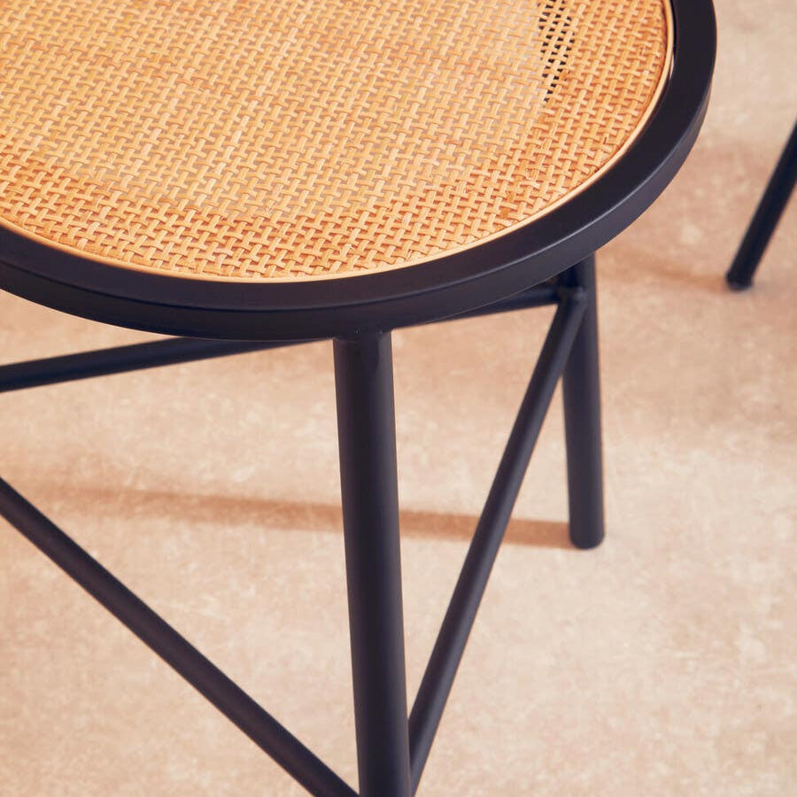 Round rattan seat stool featuring a natural woven surface and sleek black metal legs, set against a light-colored floor. The stool has a minimalist design with a sturdy frame and a simple, elegant aesthetic