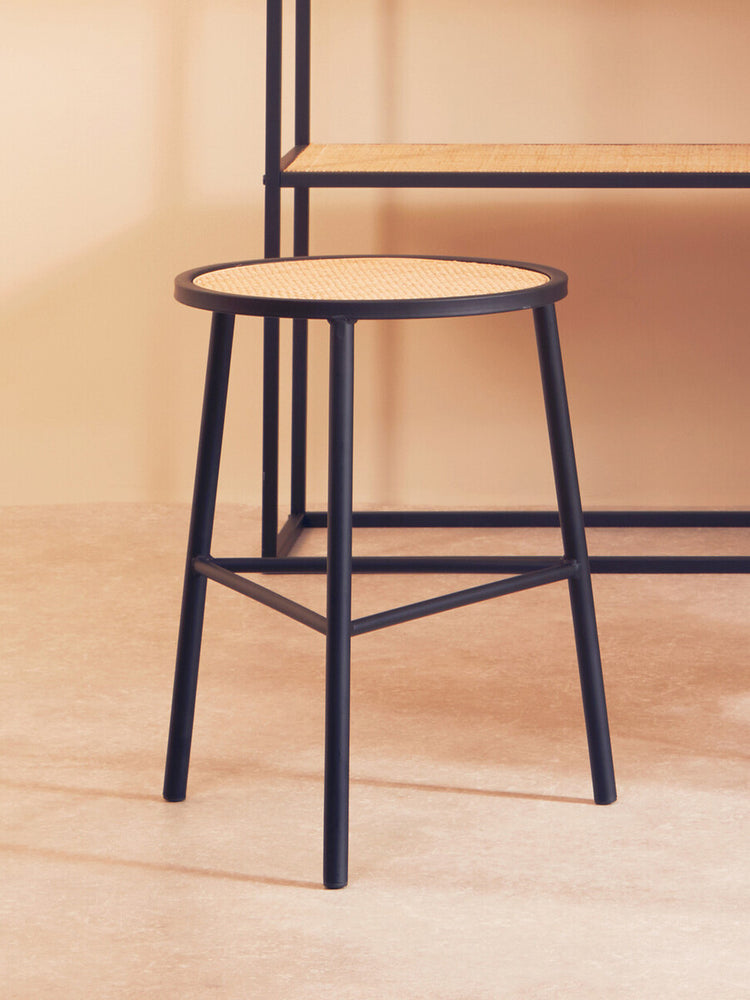 Rattan seat stool with a round top and black metal legs, positioned on a light wooden floor, with a minimalist shelving unit in the background