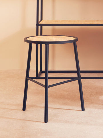 Rattan seat stool with a round top and black metal legs, positioned on a light wooden floor, with a minimalist shelving unit in the background