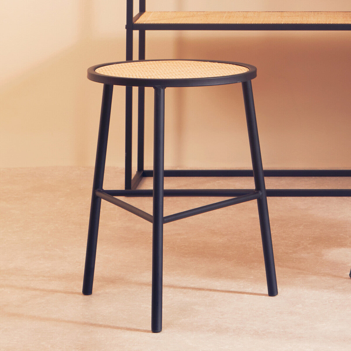 A round stool featuring a natural rattan seat and sleek black metal legs, positioned on a light-colored floor with a minimalistic backdrop