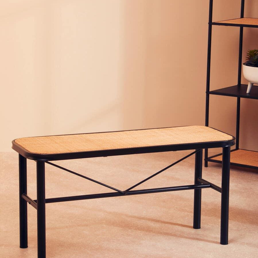 Black metal bench with woven rattan seat on beige floor near shelving unit