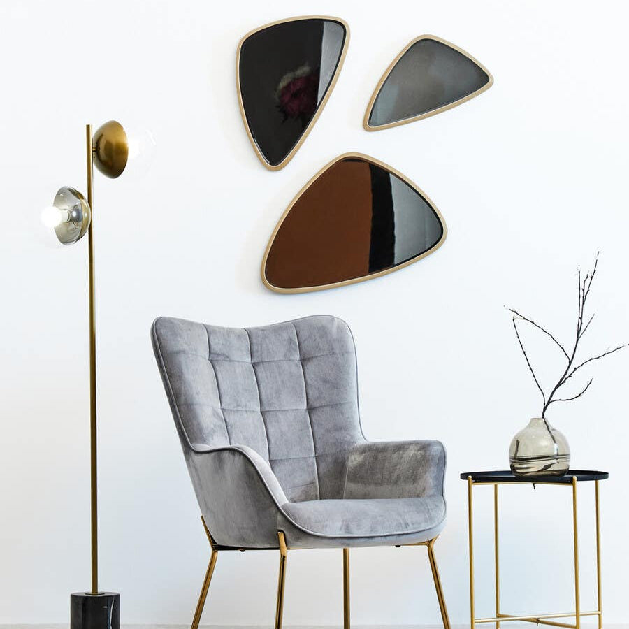 A modern black top side table with a sleek design, featuring a round surface and slender gold legs, positioned next to a stylish gray armchair. The background includes three abstract mirrors mounted on a white wall and a floor lamp with a gold shade. A decorative branch in a vase sits atop the table
