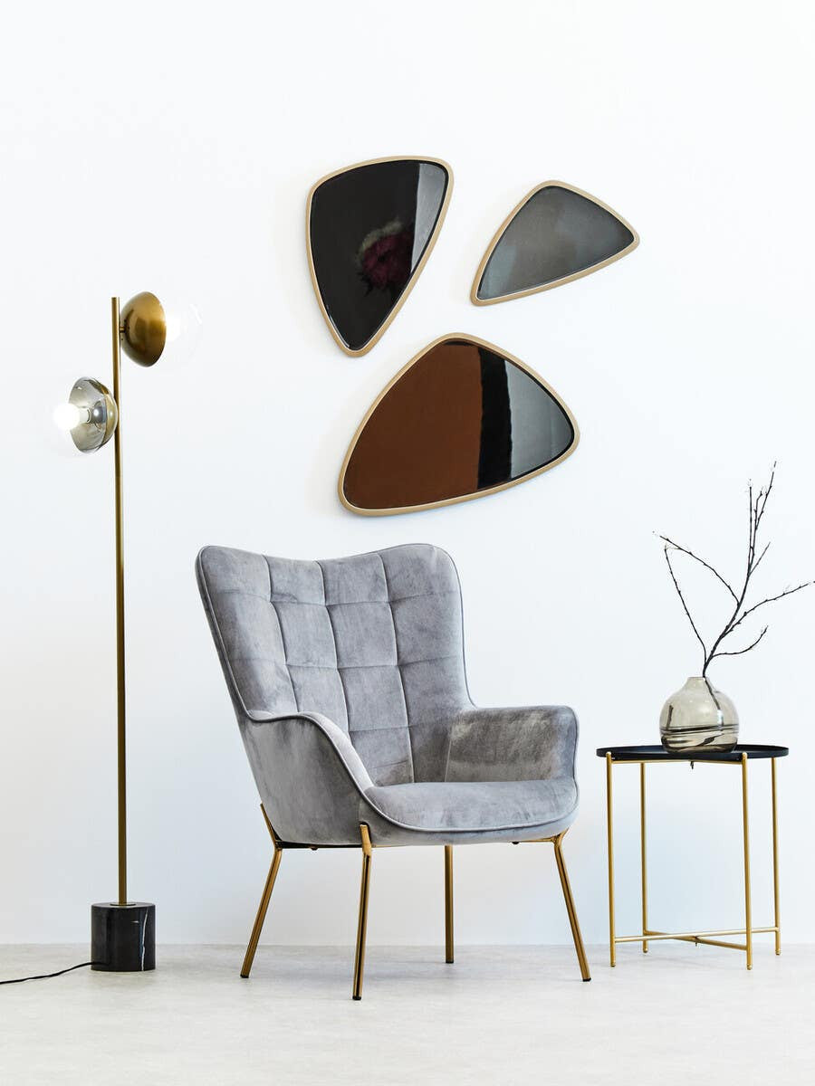 A modern black top side table with a sleek design, featuring a round surface and slender gold legs, positioned next to a stylish gray armchair. The background includes three abstract mirrors mounted on a white wall and a floor lamp with a gold shade. A decorative branch in a vase sits atop the table