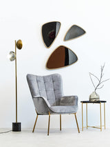 A modern black top side table with a sleek design, featuring a round surface and slender gold legs, positioned next to a stylish gray armchair. The background includes three abstract mirrors mounted on a white wall and a floor lamp with a gold shade. A decorative branch in a vase sits atop the table