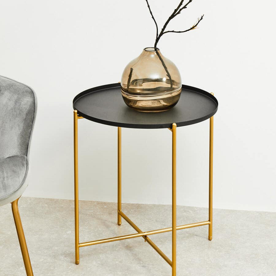 Round black top side table with gold metal legs, featuring a decorative vase with branches on top, positioned next to a gray upholstered chair on a light-colored floor