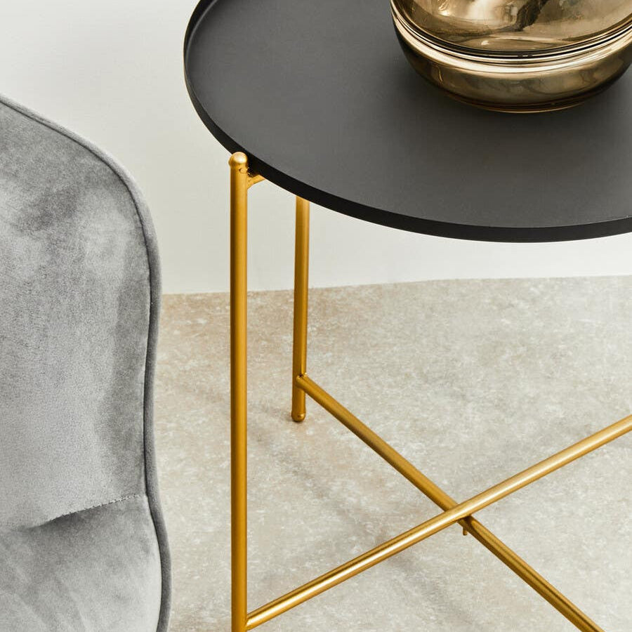 Black top side table with a round surface and gold metal legs, positioned next to a gray upholstered chair on a light-colored floor. A decorative glass vase sits on the table's surface